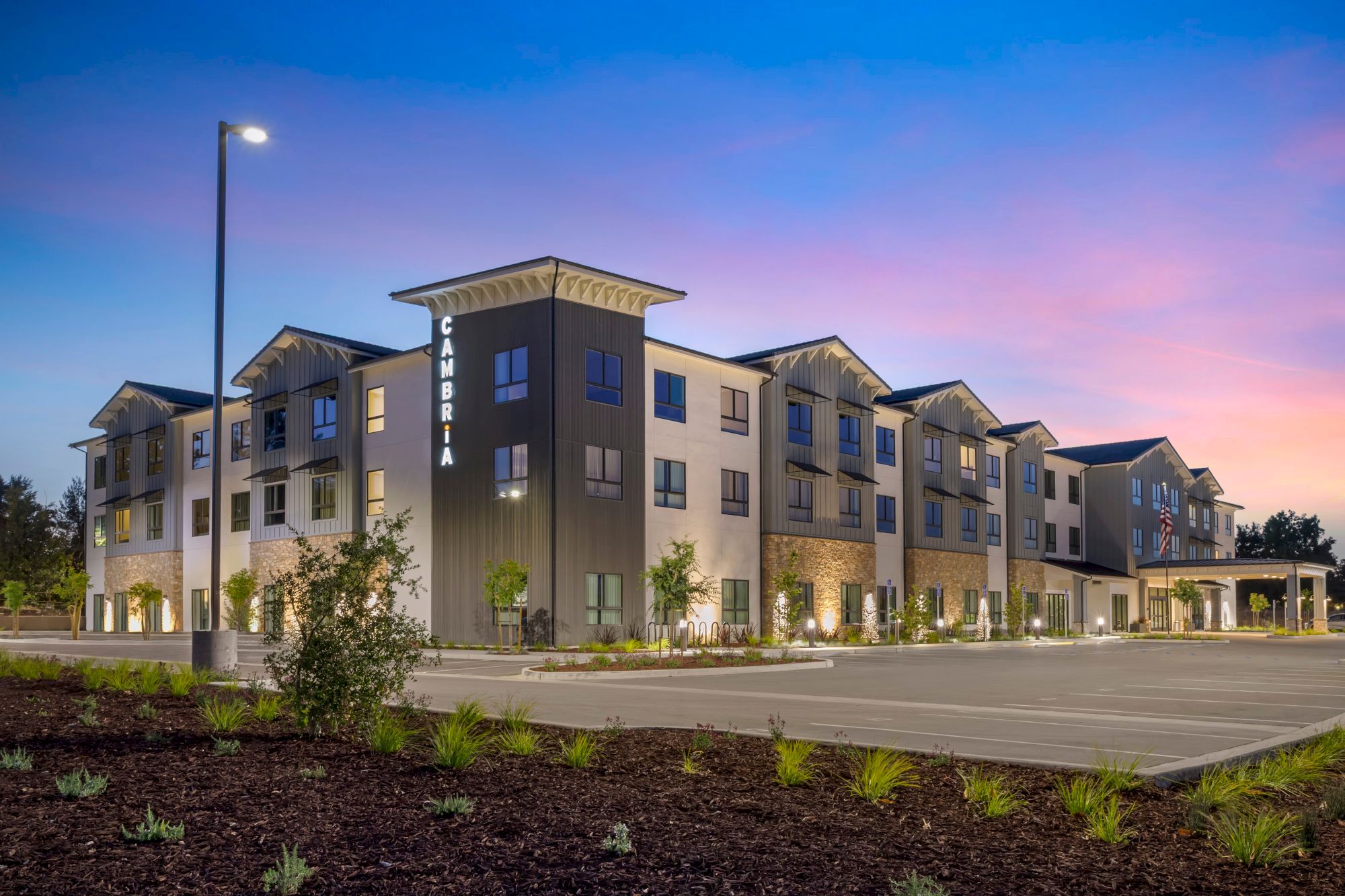 A modern apartment/hotel complex at dusk, with a tall &ldquo;CAMBRIA&rdquo; sign on the main building and streetlights along the driveway.