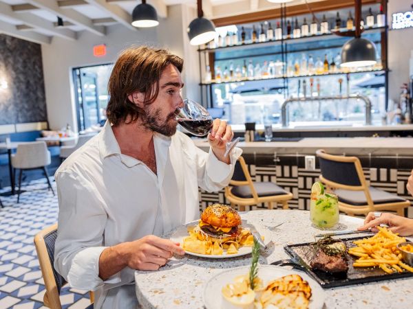 At a bar restaurant, a man in white sips red wine while eating a burger and fries in front of a grill.