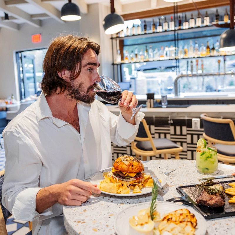 In a bar restaurant, a man in a white shirt tastes red wine beside a burger and grilled food on the table.