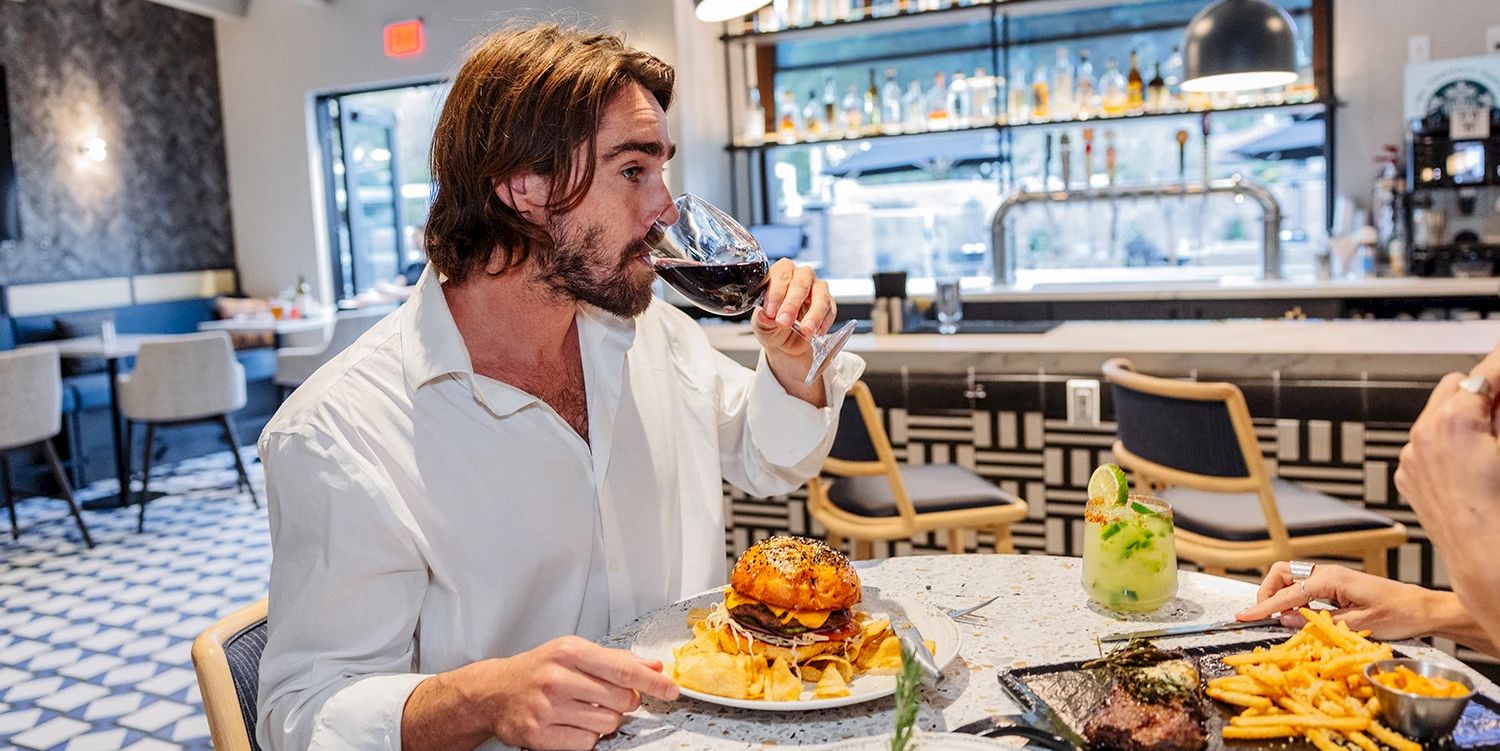 At a restaurant bar, a man sips red wine while eating a burger, fries, and steak on a shared table.