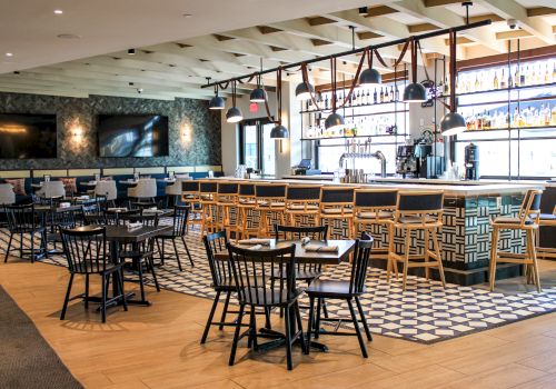 A modern bar and lounge interior with a long counter, bottle shelves, pendant lights, and dining tables and chairs.