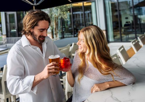 A man and woman sit on an outdoor patio under a dark umbrella, clinking drinks in the sunshine.