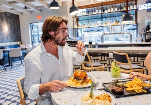 At a bar, a man in a white shirt tastes wine while eating burgers and fries in a restaurant.