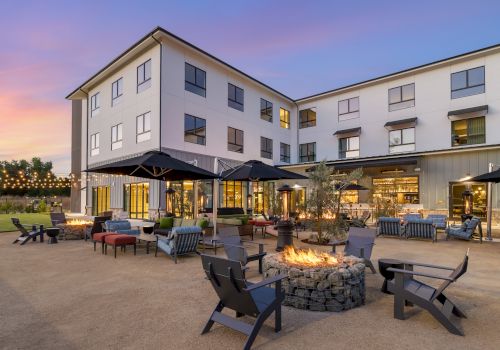 A modern apartment/hotel courtyard with outdoor seating, patio umbrellas, string lights, and a lit fire pit at dusk.