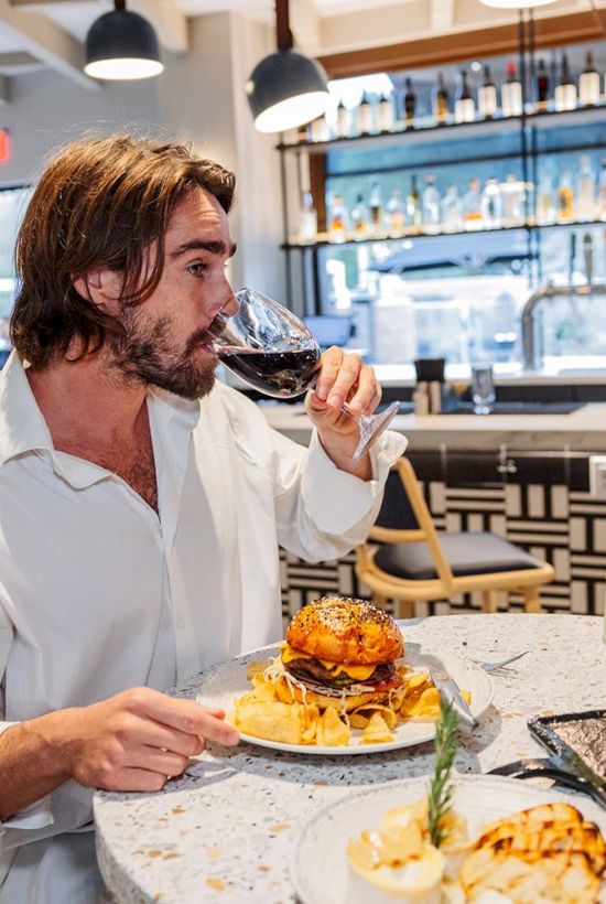 A man in a bar/restaurant tastes wine with a meal, holding a goblet while enjoying burgers and steaks.