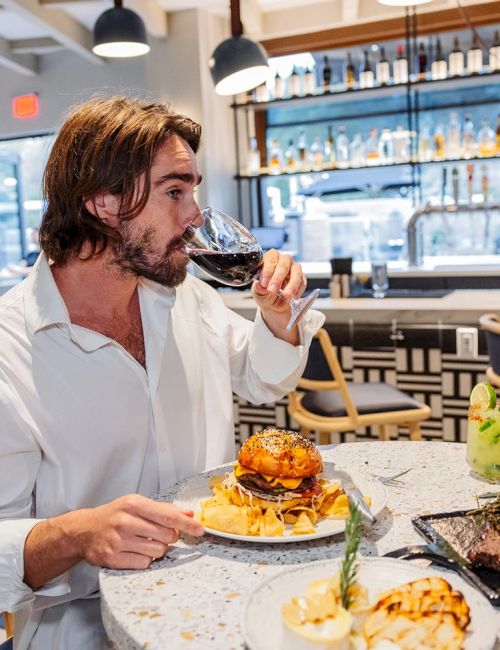A man in a bar-restaurant tastes wine while eating burgers and fries, with a bartender and beer bottles in the background.